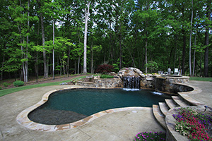 This homeowner maximized uneven elevations in their backyard by creating an elevated spillover spa finished in NPT StoneScapes Black to match the pool. Darker finishes pair well with nature-inspired elements, like this stony retaining wall and grotto.