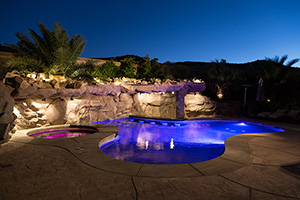 The right outdoor lighting design transforms your poolscape at night. This fun pool's colorful underwater LEDs highlight the impressive rock grotto, attached spa and in-pool stools.