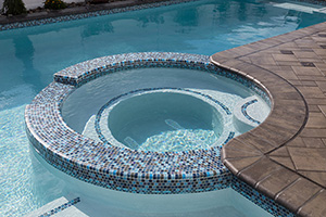 This relaxing pool design includes a raised spa covered in NPT Jules Rustic Blue Blend tile. The same tile is used on the waterline, tanning ledge, and steps for a unified design.